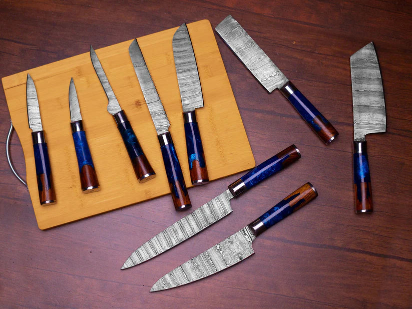 A set of kitchen knives with patterned steel blades and blue-and-brown wood handles, arranged on a wooden cutting board and a dark wood tabletop.