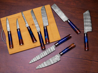 A set of kitchen knives with patterned steel blades and blue-and-brown wood handles, arranged on a wooden cutting board and a dark wood tabletop.
