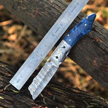A folding pocket knife with a patterned Damascus steel blade and a textured blue handle with brass pins, shown next to a metal ruler on a rustic log outdoors.