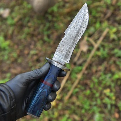 A hand wearing a black glove holds a fixed-blade knife with a patterned steel blade and a blue marbled handle with a red accent, set against a blurred outdoor background of green foliage.