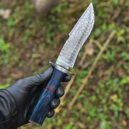 A hand wearing a black glove holds a fixed-blade knife with a patterned steel blade and a blue marbled handle with a red accent, set against a blurred outdoor background of green foliage.