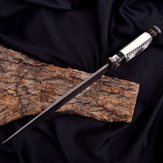 A decorative knife with a long dark blade and a white handle featuring black geometric patterns, resting diagonally on a piece of textured bark over a draped black fabric background.