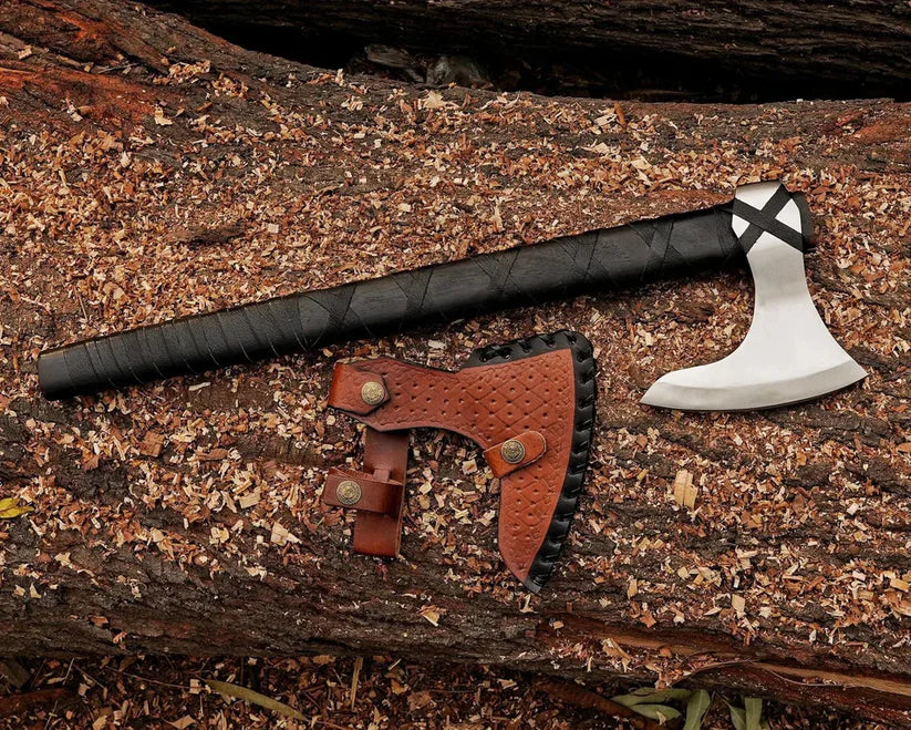 A tactical axe with a black wrapped handle and silver steel head, displayed next to its brown leather sheath on a textured log background.