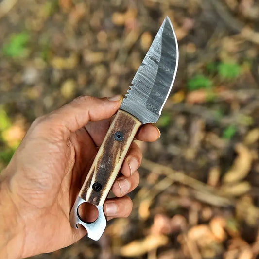 A hand holding a fixed-blade knife with a patterned steel blade and a brown antler-style handle, set against a blurred outdoor forest background.