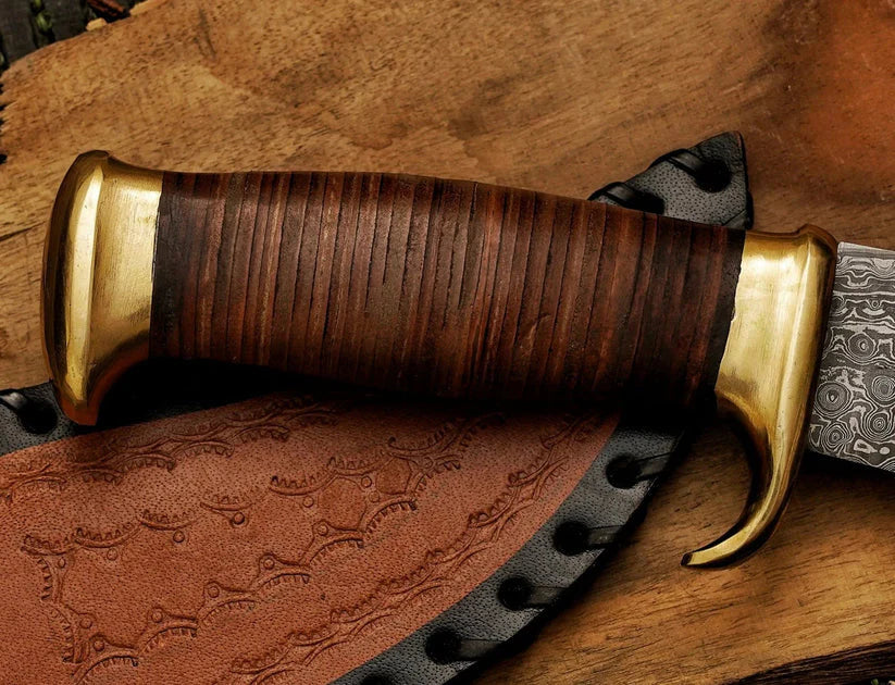 A close-up of a handcrafted knife with a dark brown wooden handle, brass guard and pommel, and a patterned steel blade, resting on a brown leather sheath over a rustic wooden surface.
