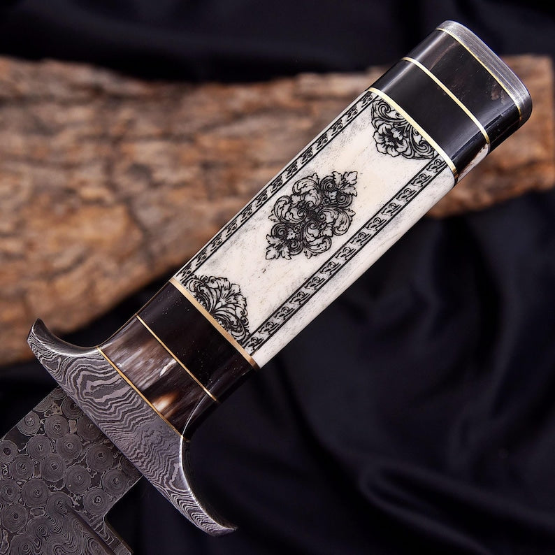 A close-up of a decorative knife handle with black floral inlay on an ivory background, dark brown sections, and gold-toned metal accents, set against a dark fabric backdrop with a piece of wood.