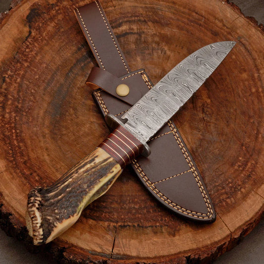 A fixed-blade knife with a patterned Damascus steel blade, a brown leather sheath, and a stag antler-style handle, displayed diagonally on a rustic wood slice background.