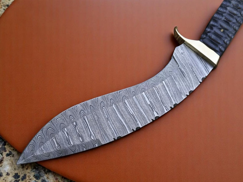 A curved Damascus steel knife with a patterned steel blade, a brass guard, and a textured black handle, resting diagonally on a brown leather surface.