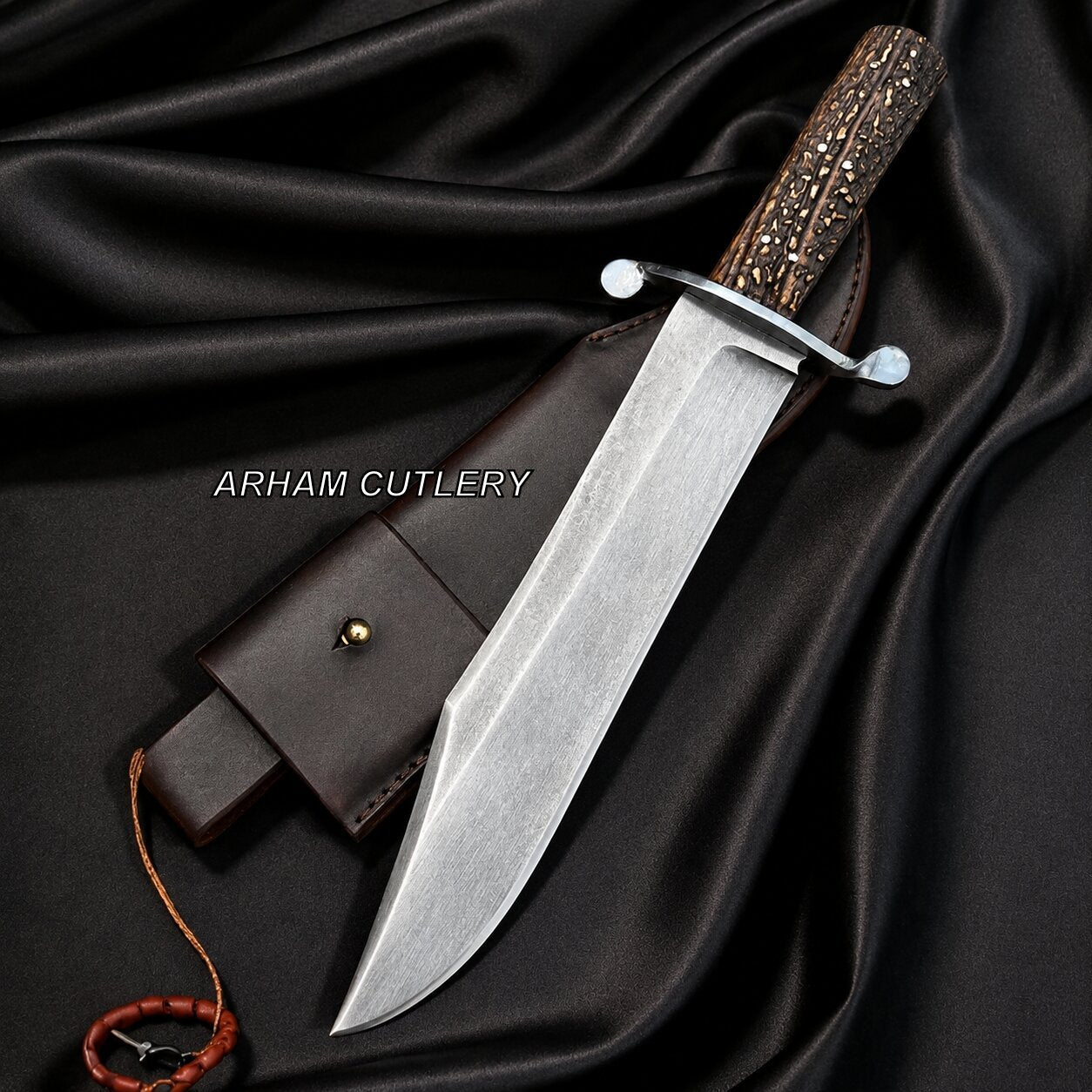 A fixed-blade knife with a silver steel blade, a textured brown handle, and a dark brown leather sheath, arranged on draped black satin fabric. The brand name 'ARHAM CUTLERY' is printed in white text across the center.