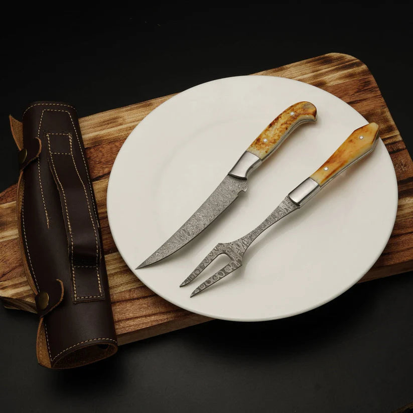 A two-piece carving set consisting of a knife and a fork with patterned steel blades and natural bone handles, displayed on a white plate over a wooden board next to its brown leather sheath.