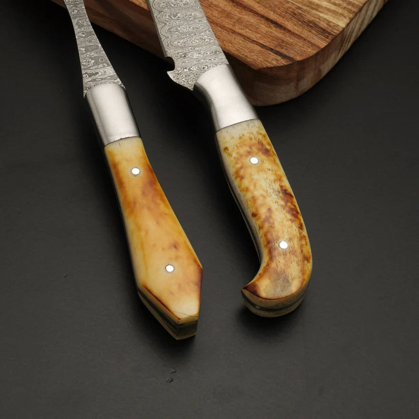 Two high-end kitchen knives with patterned steel blades and custom bone handles, displayed on a dark slate surface with a wooden cutting board in the background.