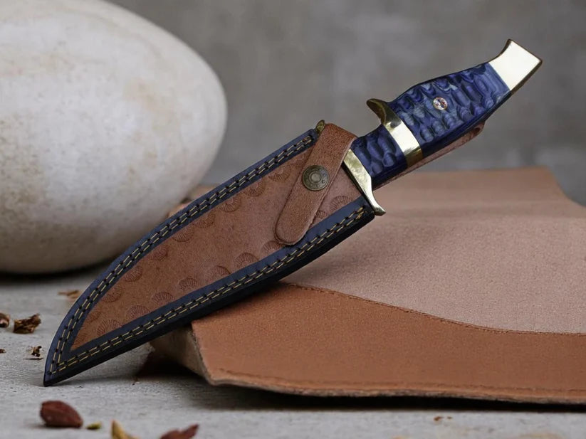 A custom knife with a blue marbled handle and brass bolsters, resting on its brown leather sheath which is placed on a piece of tan leather. The background features a neutral gray textured surface with a large white stone.