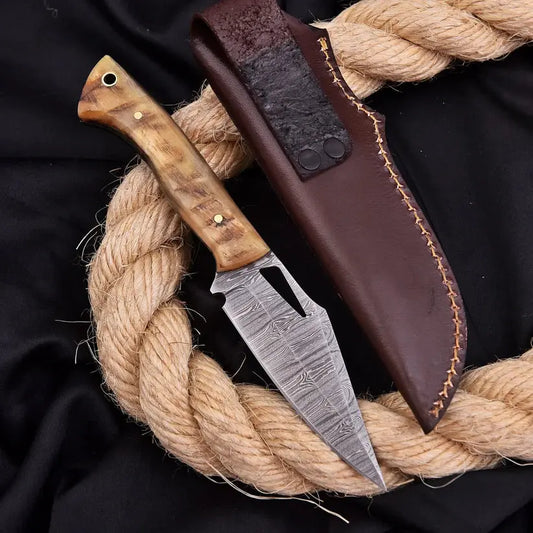 A fixed-blade knife with a patterned steel blade, a natural wood handle, and a dark brown leather sheath, arranged on a black fabric background with a thick coiled rope.