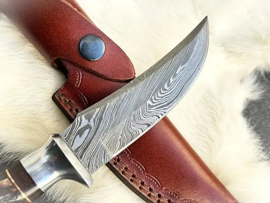 A close-up of a fixed-blade knife with a patterned Damascus-style steel blade, shown resting on a brown leather sheath over a white fur surface.