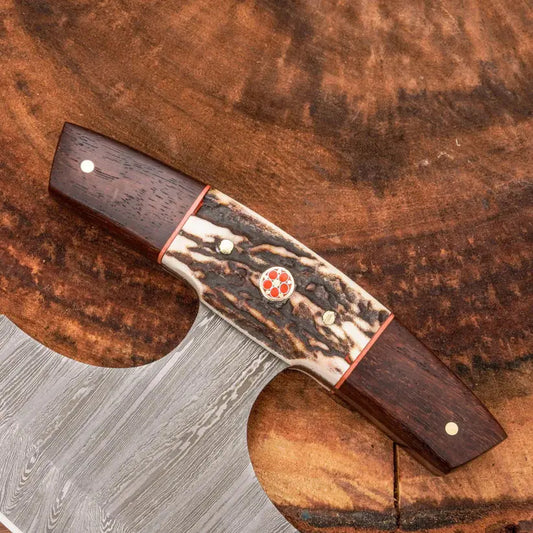 A close-up of a handcrafted kitchen knife with a Damascus steel blade and a detailed handle made of dark wood, bone, and red accents, resting on a rustic wood slice background.