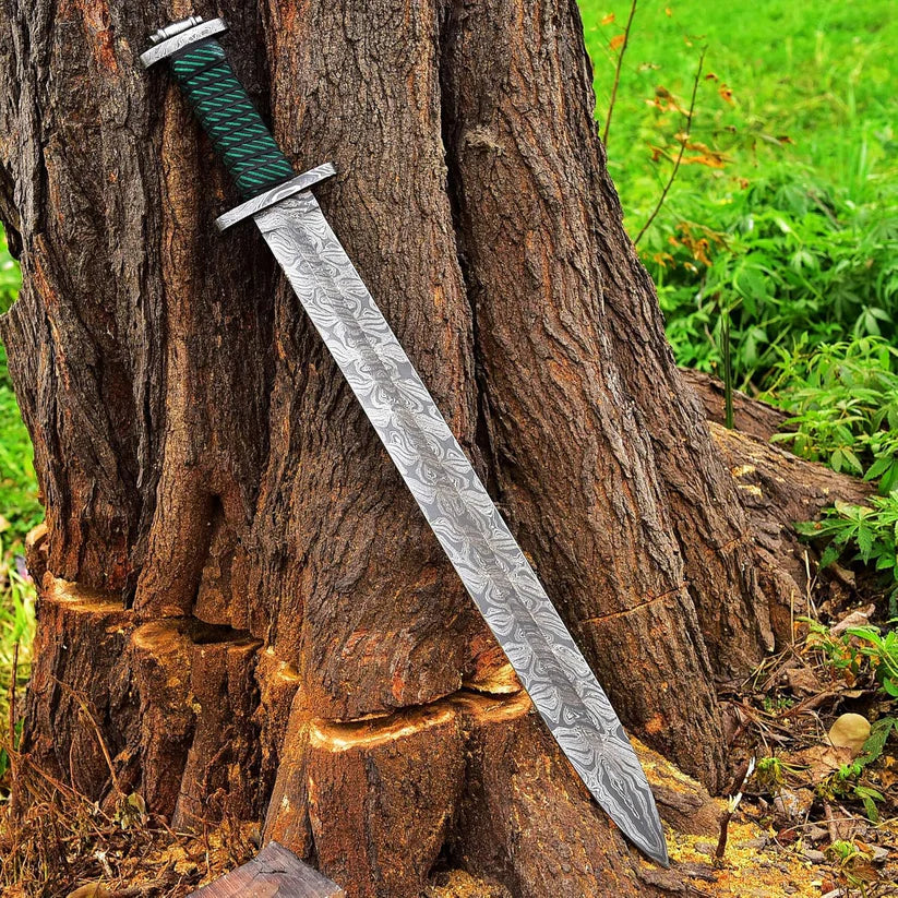A decorative sword with a patterned steel blade and a green wrapped handle, leaning against a large tree trunk outdoors.