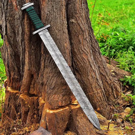 A decorative sword with a patterned steel blade and a green wrapped handle, leaning against a large tree trunk outdoors.