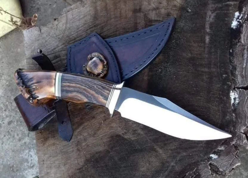 A fixed-blade knife with a polished silver blade and a carved brown handle, shown resting on a dark wooden surface next to a dark leather sheath.
