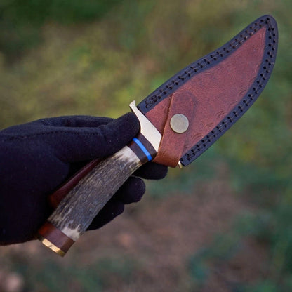 A hand wearing a black glove holds a fixed-blade knife with a bone handle, silver bolster, and blue accent band, next to a brown leather sheath with a metal snap, against a blurred outdoor background.