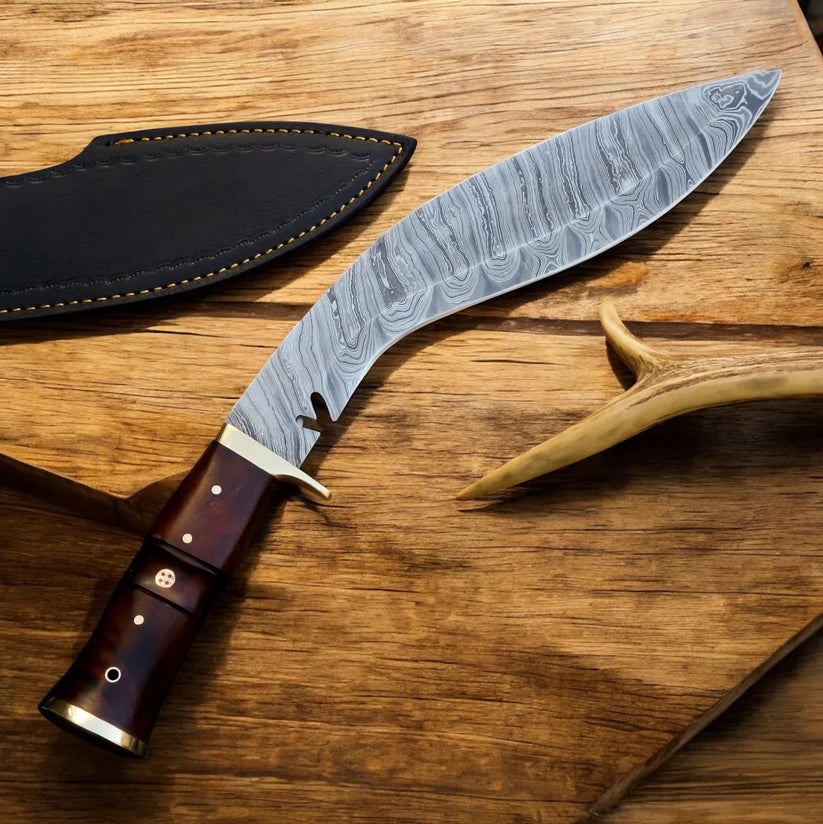 A curved Damascus steel fixed-blade knife with a dark brown wooden handle and brass pins, displayed next to its black leather sheath on a rustic wooden surface.