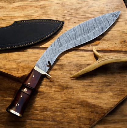 A curved Damascus steel fixed-blade knife with a dark brown wooden handle and brass pins, displayed next to its black leather sheath on a rustic wooden surface.