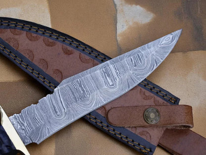 A close-up of a fixed-blade knife with a patterned Damascus-style steel blade, resting diagonally across a brown leather sheath on a tan textured surface.