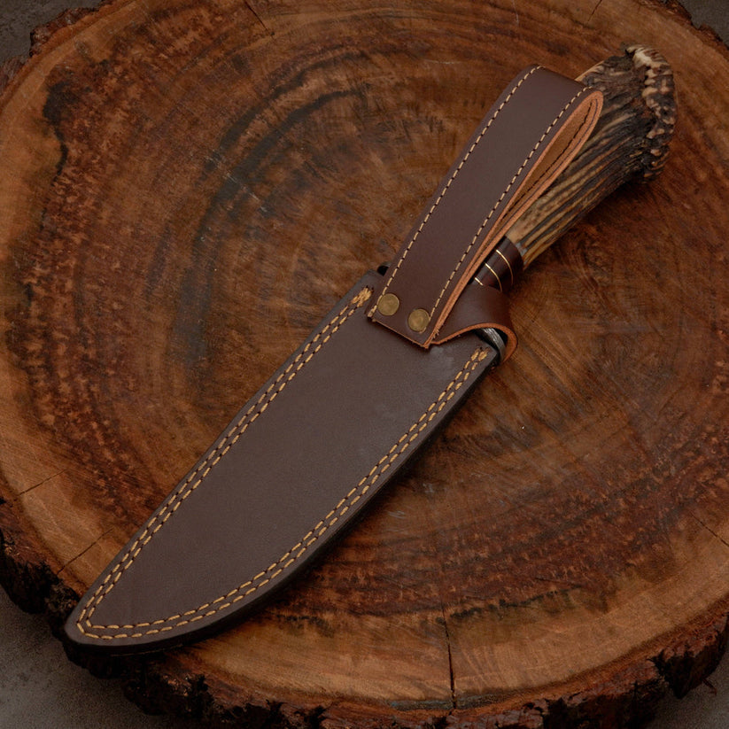 A fixed-blade knife with a natural antler-style handle and dark brown leather sheath, displayed diagonally on a rustic wood slice background.