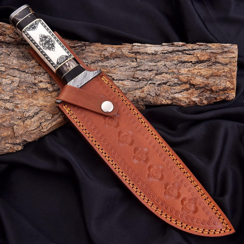 A decorative fixed-blade knife with a silver-toned engraved handle and a brown embossed leather sheath, displayed diagonally on a piece of bark over black fabric.