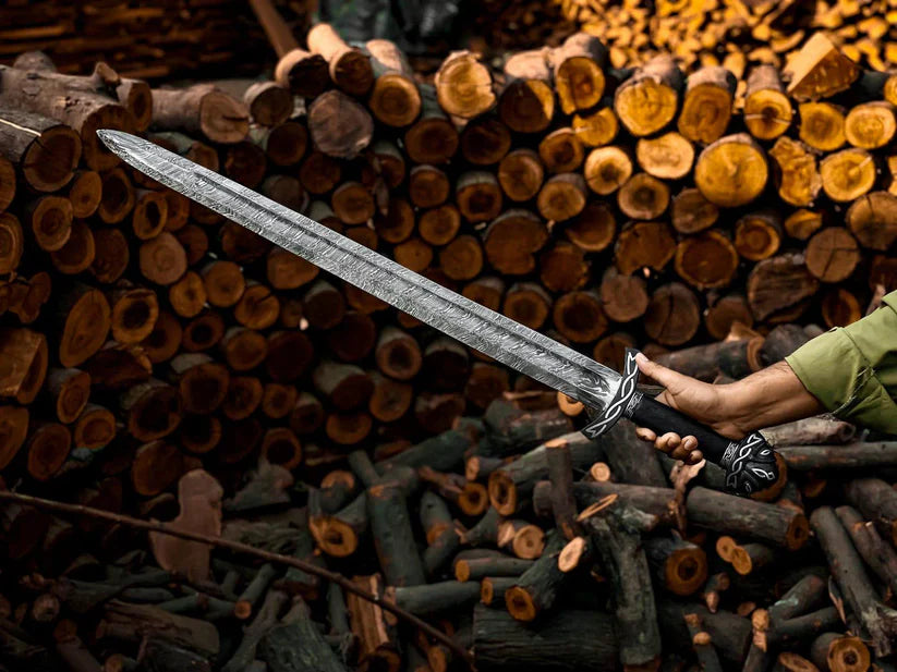 A large medieval-style sword with a textured silver blade and black hilt, held by a person's hand in front of a background of stacked firewood logs.