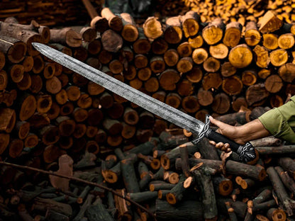 A large medieval-style sword with a textured silver blade and black hilt, held by a person's hand in front of a background of stacked firewood logs.