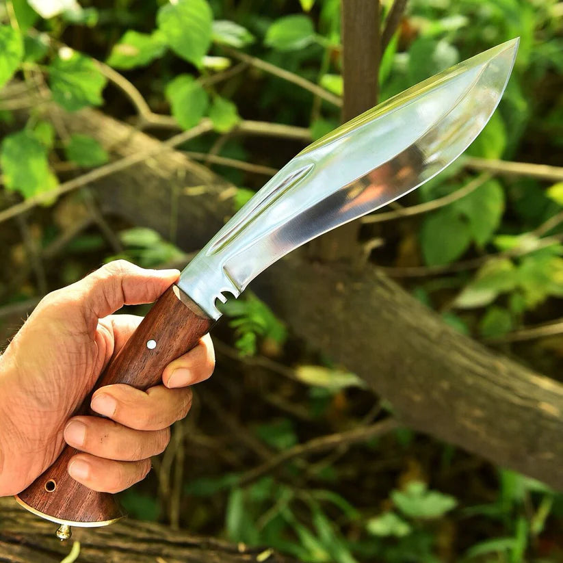 A hand holding a large machete-style knife with a polished silver blade and a brown wooden handle, set against a blurred outdoor background of green foliage and branches.