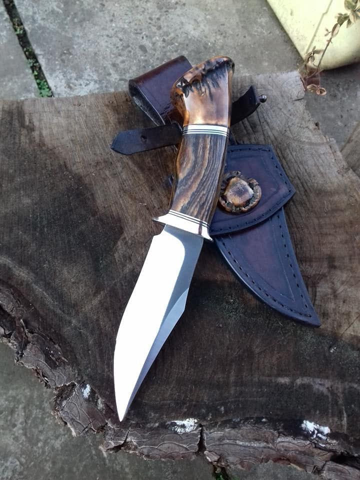 A fixed-blade knife with a polished silver blade and a brown antler-style handle, shown alongside a dark blue leather sheath on a rustic wood log outdoors.