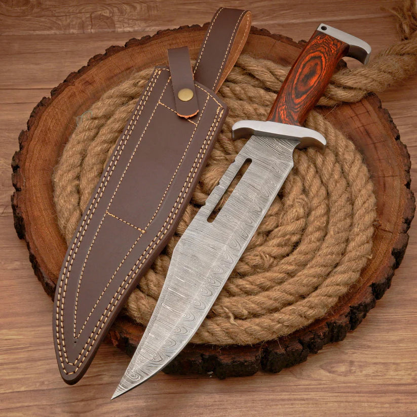 A fixed-blade knife with a patterned steel blade, silver guard, and reddish-brown wood handle, displayed next to its brown leather sheath on a rustic wood slice with coiled rope.