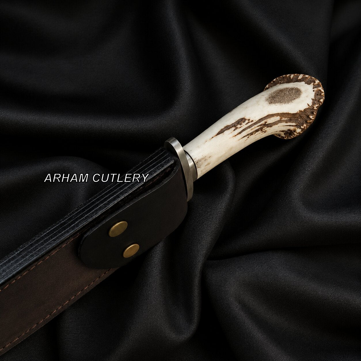 A close-up of a handcrafted knife with an antler-style handle and a dark brown leather sheath, displayed on draped black satin fabric. The brand name 'ARHAM CUTLERY' is printed in white text on the sheath.