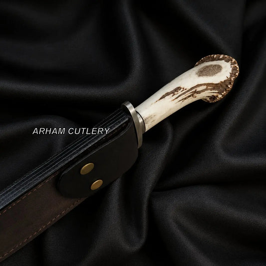 A close-up of a handcrafted knife with an antler-style handle and a dark brown leather sheath, displayed on draped black satin fabric. The brand name 'ARHAM CUTLERY' is printed in white text on the sheath.