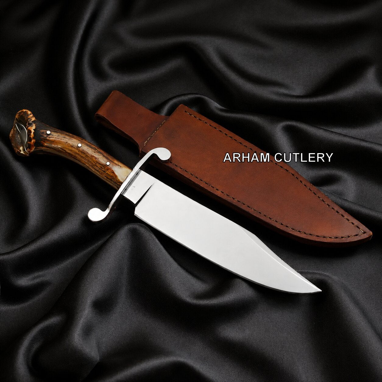 A fixed-blade knife with a polished silver blade, a brown wood-toned handle, and a brown leather sheath, arranged on draped black satin fabric. The text 'ARHAM CUTLERY' is printed on the sheath.
