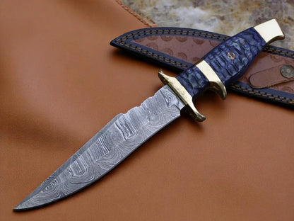 A fixed-blade knife with a patterned steel blade, a blue scaled handle, and brass guard, displayed diagonally on a brown leather sheath over a brown leather surface.