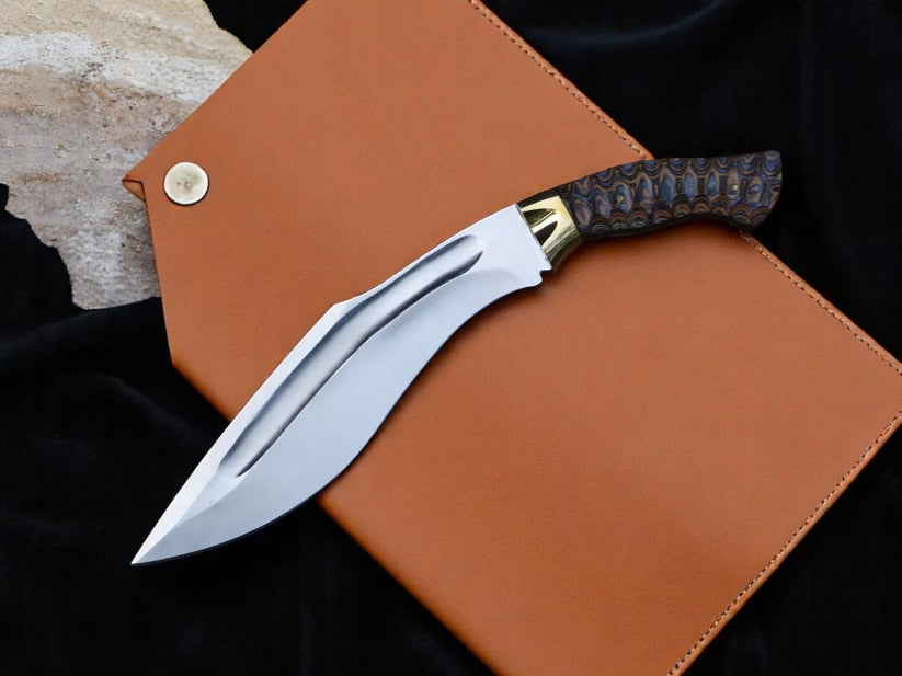A curved fixed-blade knife with a polished silver blade, gold-toned bolster, and dark patterned handle, displayed diagonally on a brown leather sheath over a black fabric background.