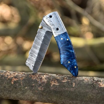A folding pocket knife with a Damascus steel blade and a blue marbled handle, resting on a tree branch with a blurred forest background.