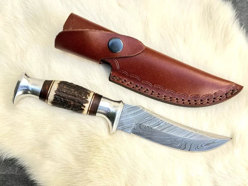 A fixed-blade knife with a patterned steel blade, a horn and wood handle, and a brown leather sheath, displayed on a white fur background.