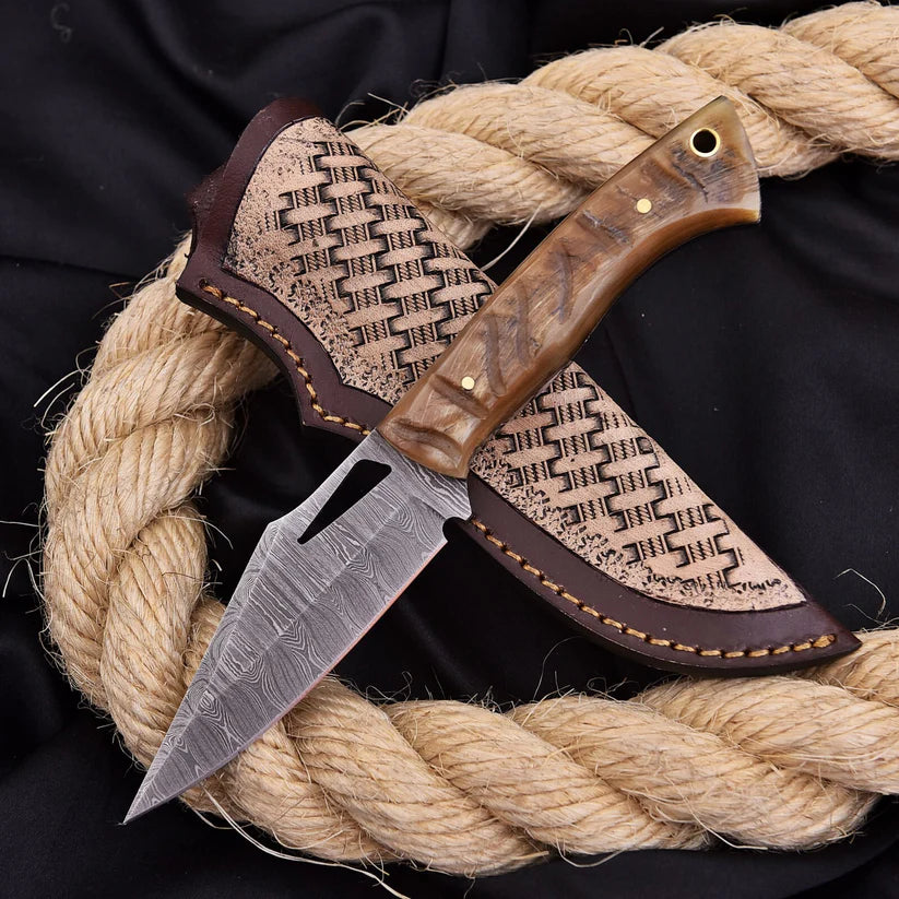 A fixed-blade knife with a patterned steel blade and a brown wood-grain handle, shown partially sheathed in a matching brown leather sheath. The knife is arranged on a thick coiled natural rope over a black fabric background.