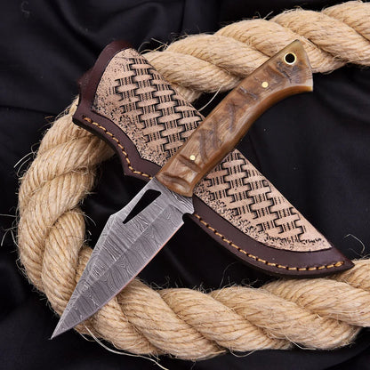 A fixed-blade knife with a patterned steel blade and a brown wood-grain handle, shown partially sheathed in a matching brown leather sheath. The knife is arranged on a thick coiled natural rope over a black fabric background.