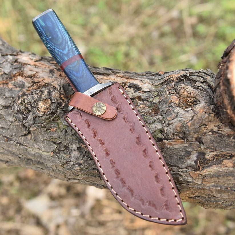 A fixed-blade knife with a blue marbled handle and a brown leather sheath, resting on a textured tree branch outdoors.