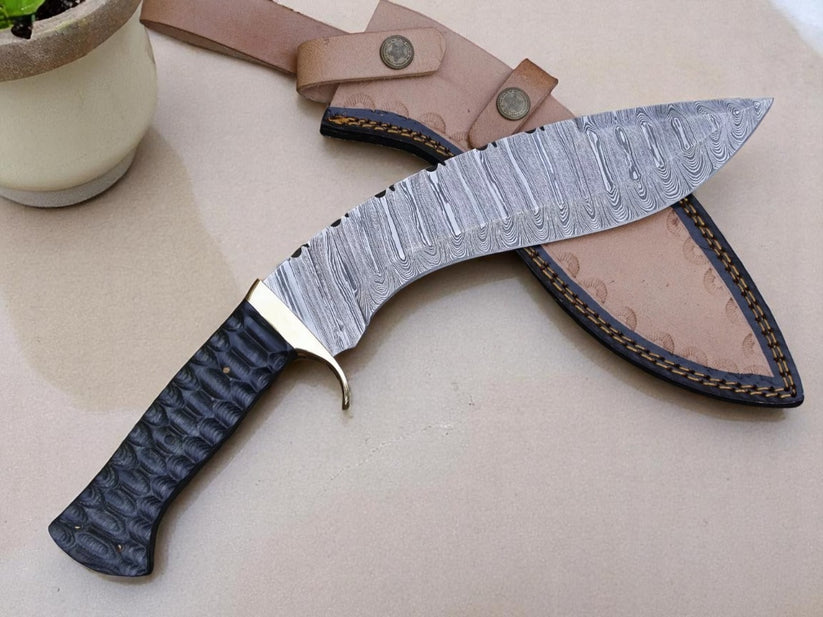 A curved fixed-blade knife with a patterned steel blade, textured black handle, and a tan leather sheath, displayed on a beige surface with a small potted plant in the corner.