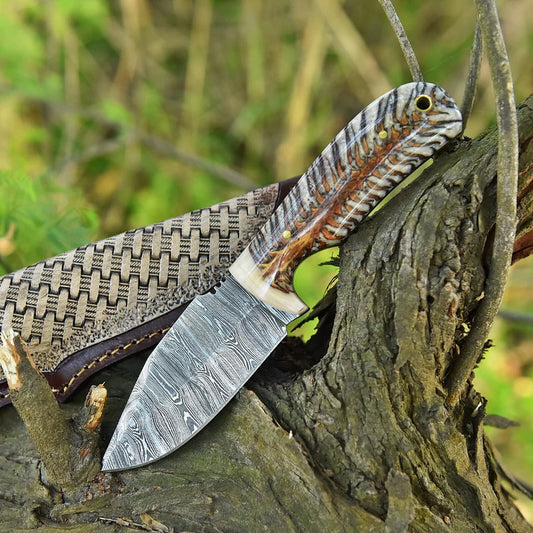 A fixed-blade knife with a patterned Damascus-style steel blade and a textured handle, resting on a tree trunk outdoors with a woven sheath behind it.