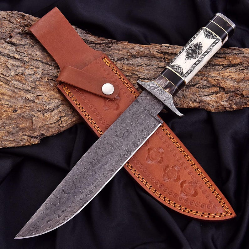 A fixed-blade knife with a patterned steel blade, black and white handle, and a brown leather sheath, displayed on a piece of wood over black fabric.
