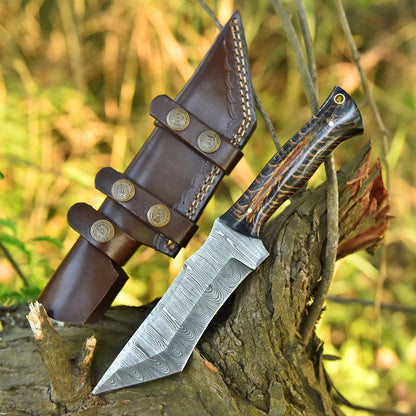 A fixed-blade knife with a patterned steel blade and a carved handle, resting on a tree stump outdoors next to a brown leather sheath with brass snaps.
