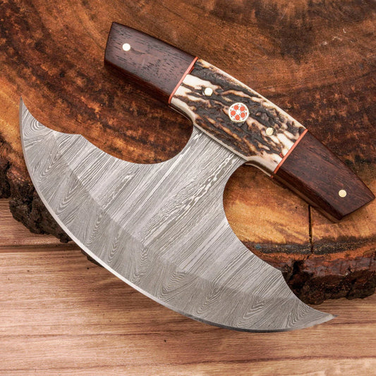 A handcrafted axe with a Damascus steel blade and a decorative handle made of dark wood and patterned bone, displayed on a rustic wood slice background.