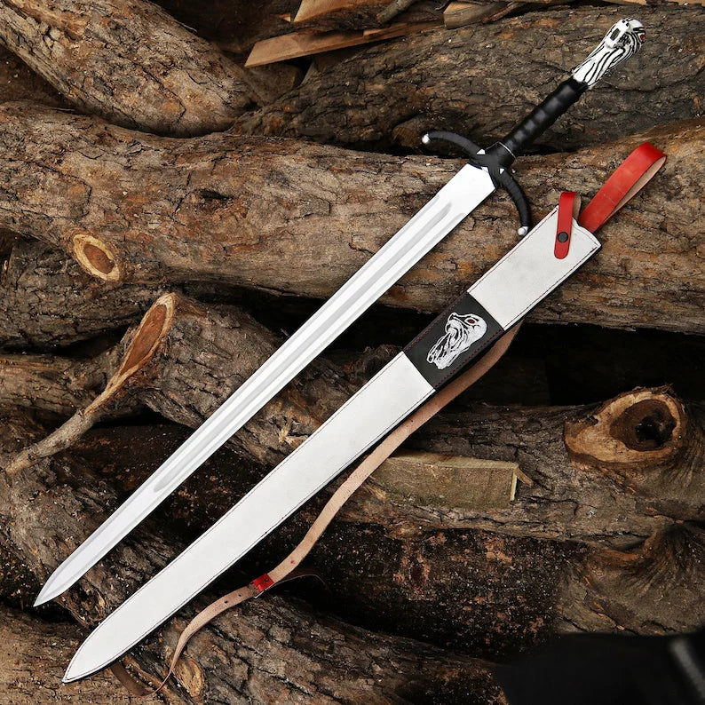 A long sword with a silver blade, black handle, and decorative silver pommel lies diagonally on stacked logs, alongside its matching sheath with a red strap and a small red tag.