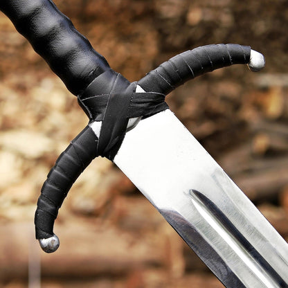 A close-up of a medieval-style sword with a black leather-wrapped hilt and a silver steel blade, set against a blurred outdoor background of brown leaves and logs.